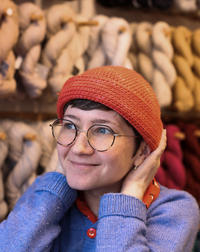 Cleo, a white non-binary person, wears an orange crocheted hat with the brim rolled up. They have their hands touching the nape of their neck and look happy.